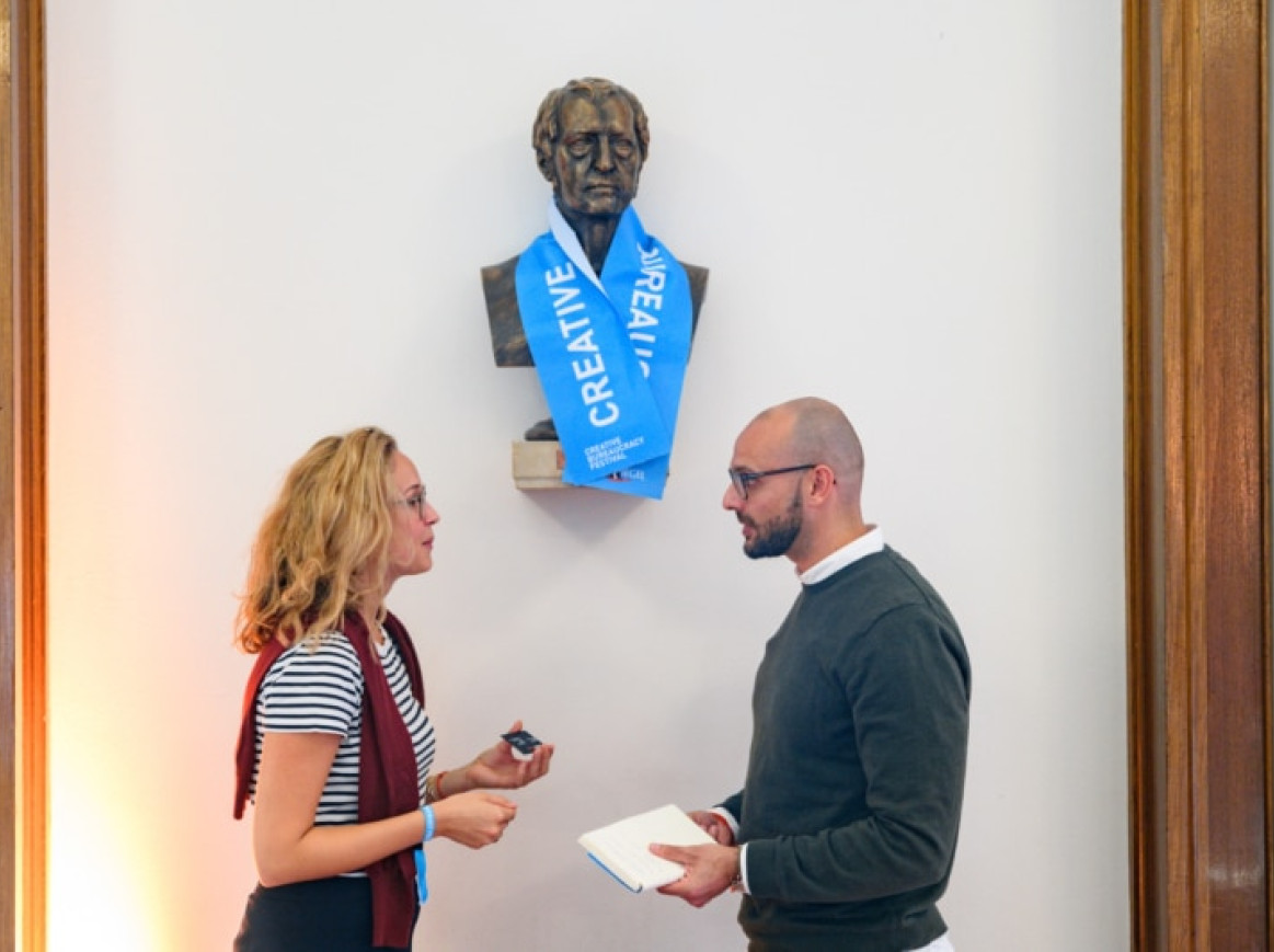 Mann und Frau sprechen vor einer Steinstatue mit Merchandise der kreativen Bürokratie