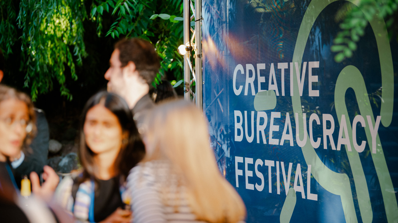 people at the festival area outside standing and talking infront of a big poster of the festival 