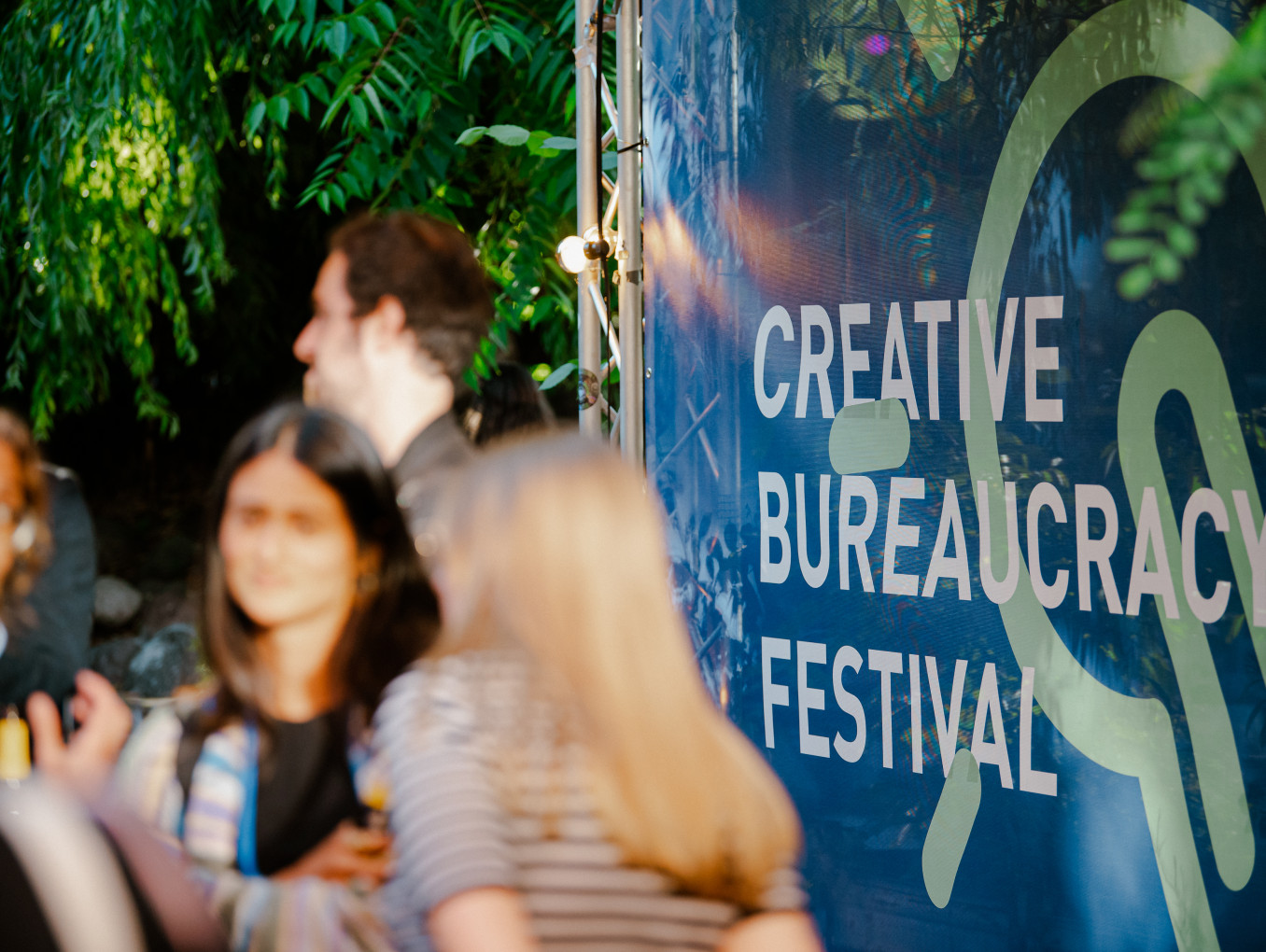 people at the festival area outside standing and talking infront of a big poster of the festival