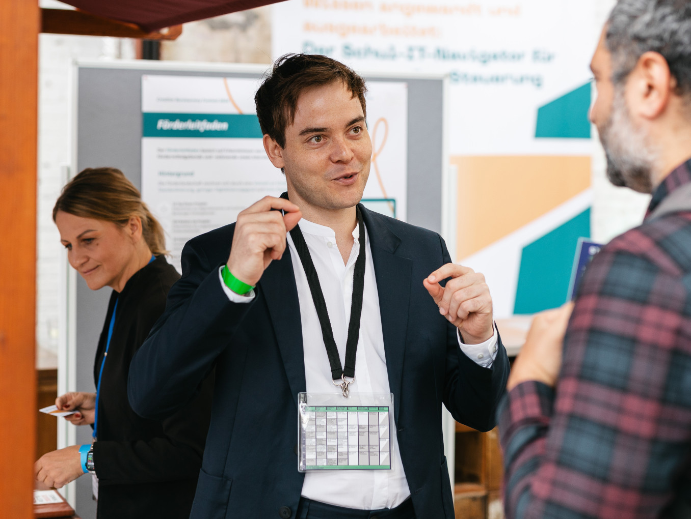 Mann erklärt einem anderen Mann etwas, während sie neben einem Stand stehen, und eine Frau hinter ihnen schaut auf den Stand
