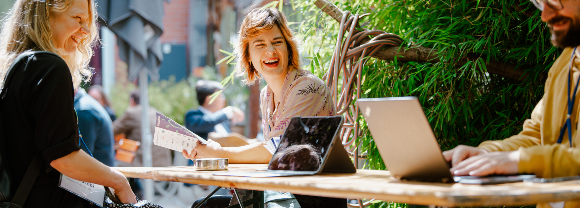 Drei Personen sitzen an einem Tisch, lachen und schauen auf einen Laptop