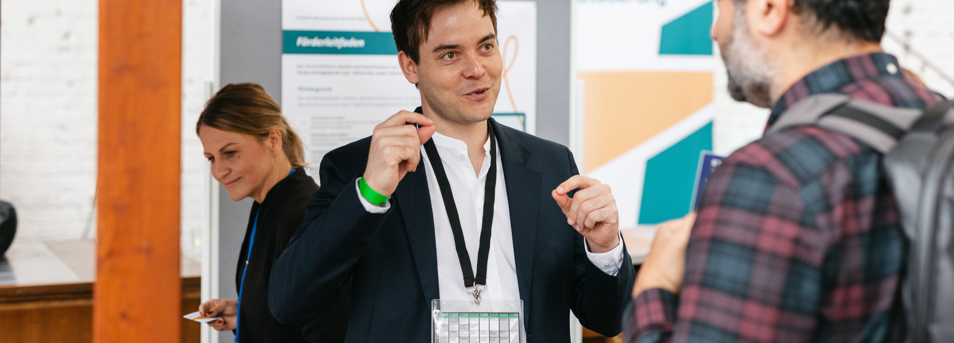 Mann erklärt einem anderen Mann etwas, während sie neben einem Stand stehen, und eine Frau hinter ihnen schaut auf den Stand